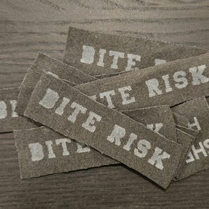 May include: A collection of rectangular olive green fabric patches featuring the text BITE RISK printed in a light gray, block-style font. The patches are layered on top of one another, showing a woven texture and frayed edges on the fabric strips.