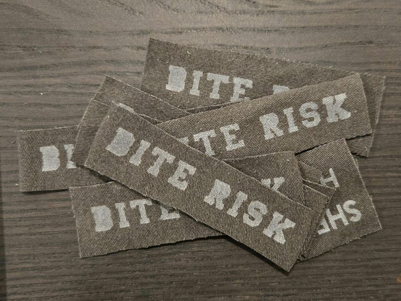 May include: A collection of rectangular olive green fabric patches featuring the text BITE RISK printed in a light gray, block-style font. The patches are layered on top of one another, showing a woven texture and frayed edges on the fabric strips.