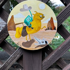 May include: A hand-painted round canvas featuring a cartoon dog wearing a cowboy hat and riding a green robot. The background is a desert scene with mountains and clouds. The text "B.M.C." is on the robot's chest.