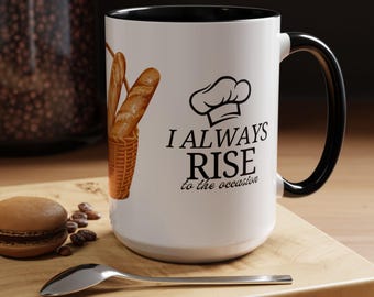 Könnte beinhalten: Weißer Keramikbecher mit schwarzem Innenraum und Henkel. Der Becher zeigt einen Brotkorb und den Text "I ALWAYS RISE to the occasion" mit einer Kochmützen-Grafik. Ein Macaron und ein Löffel liegen auf einer Holzoberfläche.
