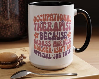 May include: White ceramic mug with a black handle and rim. The mug features the text "OCCUPATIONAL THERAPIST BECAUSE BADASS MIRACLE WORKER ISN'T AN OFFICIAL JOB TITLE" in retro-style lettering. The mug sits on a wooden surface.