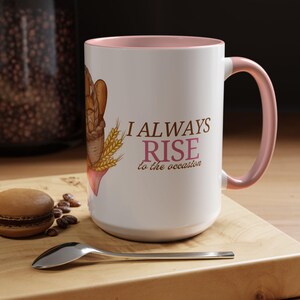 Peut inclure: Mug en céramique blanche avec intérieur et anse roses. L'illustration représente du pain et le texte "I ALWAYS RISE to the occasion" en marron et rose. Un macaron, des grains de café et une cuillère sont sur une surface en bois.