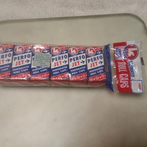 May include: A package of Perfo Jet perforated roll caps. The packaging is red and white with the words "PERFO JET" and "PERFORATED ROLL CAPS" printed on it. The package also includes a small, separate packet of roll caps.