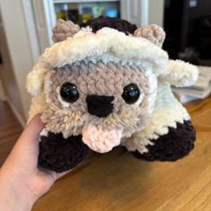 May include: A crocheted plush toy shaped like a sheep with brown and white fur. The sheep has large black eyes and a pink tongue sticking out.