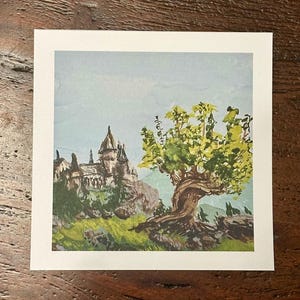 May include: A square art print featuring a watercolor-style illustration of a stone castle perched on a rocky hill next to a large, twisted tree with vibrant green and yellow leaves against a pale blue sky, set within a white border.