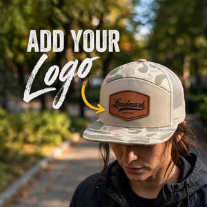 May include: A beige and light green camouflage trucker hat featuring a brown leather patch on the front panel. The patch displays the text Landmark and Achievement in black script and print. The text ADD YOUR Logo is visible in the image.