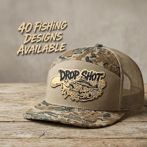 May include: A camouflage and tan trucker hat featuring a patch with a fish graphic and the text "DROP SHOT". The text "40 FISHING DESIGNS AVAILABLE" is visible above the hat. The hat has a mesh back and a solid front panel with a flat brim.