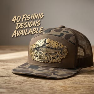 May include: A brown camouflage trucker hat with a mesh back and a gold-toned patch on the front. The patch features a fish illustration, a crescent moon, stars, and the text "NIGHT BITE". The text "40 FISHING DESIGNS AVAILABLE" appears above.