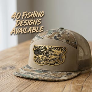 May include: A tan and camouflage trucker hat featuring a patch with the text AMERICAN WHISKERS and a graphic of a catfish wearing a bandana in front of an American flag. The text 40 FISHING DESIGNS AVAILABLE is displayed above the hat.