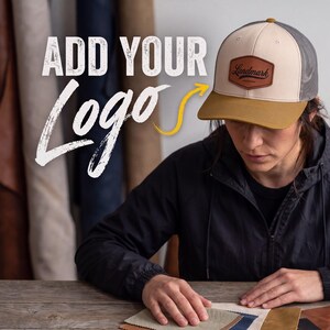 May include: A trucker hat with a cream-coloured front panel, a brown brim, and grey mesh back panels. A brown leather patch is centred on the front with the text Landmark. The text ADD YOUR Logo is visible to the left of the hat with a yellow arrow.