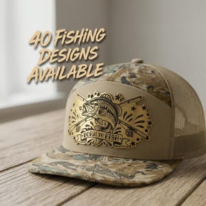 May include: A camouflage trucker hat with a tan front panel featuring a gold-toned metal patch. The patch displays a fish, two crossed fishing rods, stars, and the text "BORN TO FISH". The text "40 FISHING DESIGNS AVAILABLE" appears above the hat.