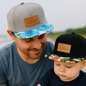 Könnte beinhalten: Zwei Baseballkappen, eine graue und eine schwarze, mit Lederaufnähern. Auf der grauen Kappe steht "Dad", auf der schwarzen "Williams". Beide haben blumige Krempen. Die Kappen werden von einem Mann und einem Kind getragen.