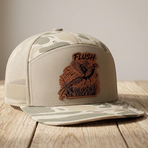 Op de afbeelding: Een beige en lichtbruine camouflage truckerpet met een bruine leren patch op het voorpaneel. De patch toont een zwarte afbeelding van een fazant die opvliegt uit hoog gras, met het woord FLUSH in een vetgedrukt lettertype erboven.