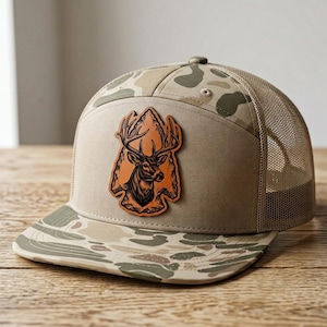 May include: A tan and olive green camouflage trucker cap featuring a brown leather patch on the front panel. The patch displays an engraved illustration of a deer head centred within an arrowhead shape. The cap has a mesh back and flat brim.