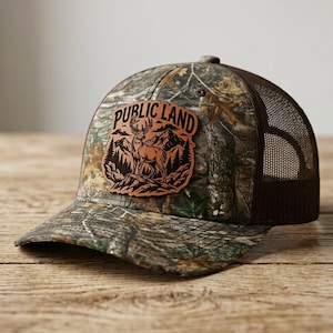 Könnte beinhalten: Eine Camouflage-Baseballkappe mit braunem Netzrücken und einem braunen Lederaufnäher auf der Vorderseite. Der Aufnäher zeigt eine schwarze Grafik eines Hirsches im Wald mit Bergen im Hintergrund und dem Text PUBLIC LAND in einem Bogen darüber.