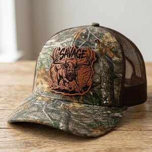 Könnte beinhalten: Eine Camouflage-Trucker-Cap mit einem braunen Lederaufnäher auf der Vorderseite. Der Aufnäher zeigt eine eingravierte Abbildung eines Hirsches mit großem Geweih und das Wort SAVAGE in einer fetten, stilisierten Schrift über dem Tier.