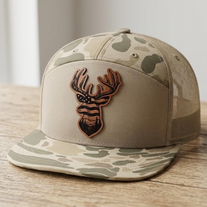 May include: A tan and beige camouflage trucker hat featuring a flat brim and a mesh back. The front panel displays a brown leather-like patch shaped like a deer head, which contains an engraved design of the American flag and a forest landscape.