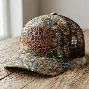 May include: A camouflage baseball cap with a brown mesh back and a brown leather patch on the front. The patch features an antler design and the text I LIKE BIG BUCKS AND I CANNOT LIE engraved in black lettering on the tan leather surface.