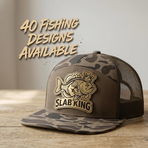 May include: A brown camouflage trucker hat featuring a gold-toned patch on the front panel. The patch displays a fish wearing a crown with the text SLAB KING below it. The text 40 FISHING DESIGNS AVAILABLE is positioned above the hat in brown.