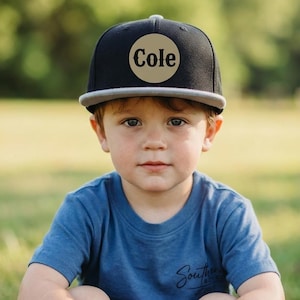 Può includere: Un cappellino da baseball nero con visiera piatta grigia e una toppa circolare color marrone chiaro sul davanti. La toppa presenta il nome Cole stampato in un carattere serif nero in grassetto. Il cappellino ha una corona strutturata e un bottone.
