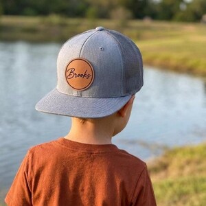 Peut inclure: Une casquette de baseball grise avec une visière plate et un panneau arrière en maille. L'avant présente un écusson circulaire en cuir beige avec le nom Brooks gravé dans une police manuscrite sombre. La casquette a une calotte structurée.