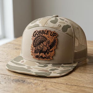 May include: A tan trucker hat featuring a camouflage pattern on the brim and top panels. The front panel displays a tan leather-like patch with a black engraved illustration of a turkey and the text GOBBLE UP in a bold, stylized font.