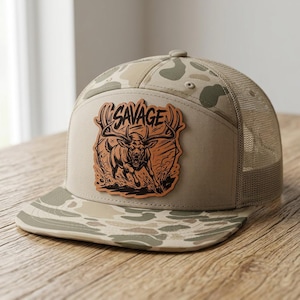 May include: A tan and beige camouflage trucker hat featuring a leather-like patch on the front. The patch displays an illustration of a charging deer with large antlers and the word SAVAGE printed in a bold, stylized font above the animal.