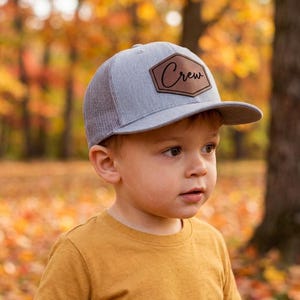 May include: A heather gray trucker hat with a mesh back and a stiff brim. Centered on the front panel is a brown, hexagon-shaped leather patch featuring the word Crew in a black, cursive script font. The hat has a structured crown and stitching.