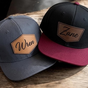 May include: Two baseball caps with flat brims and structured crowns. One cap is dark grey with a tan leather-like patch featuring the name Wren. The other cap is black with a maroon brim and a tan leather-like patch featuring the name Zane.