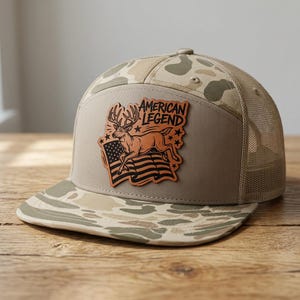 Könnte beinhalten: Eine beigefarbene Camouflage-Trucker-Cap mit einem Lederaufnäher auf der Vorderseite. Der Aufnäher zeigt einen springenden Hirsch über einer amerikanischen Flagge mit dem Schriftzug AMERICAN LEGEND und mehreren kleinen Sternen.