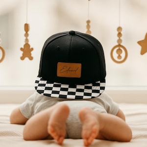 May include: A black baseball cap with a chequered brim and a brown leather patch with the name "Edward" in script. The cap is worn by a baby lying on a bed. Wooden ornaments hang in the background.