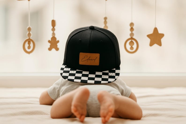 May include: A black baseball cap with a chequered brim and a brown leather patch with the name "Edward" in script. The cap is worn by a baby lying on a bed. Wooden ornaments hang in the background.