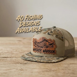 May include: A camouflage trucker hat with a tan front panel featuring a leather patch with a fish graphic and the text "WHISKER WARRIOR". The text "40 FISHING DESIGNS AVAILABLE" is displayed above the hat in a tan, stylized font.