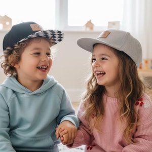 May include: Two children wearing caps and smiling. One child wears a black cap with a chequered brim, the other a grey cap. The children are wearing a light blue hoodie and a pink sweater, respectively. They are holding hands.