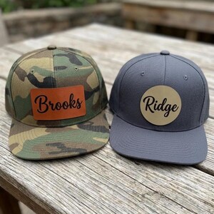 May include: Two baseball caps, one with a green, brown, and black camouflage pattern featuring a rectangular brown leather patch with the word Brooks, and one solid charcoal gray cap with a circular tan leather patch with the word Ridge.