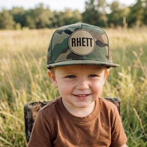 Puede incluir: Una gorra de béisbol de camuflaje con un parche redondo de color tostado en el panel frontal. El parche tiene impreso el nombre RHETT en letras mayúsculas negras y negritas. La gorra tiene una corona estructurada y una visera plana.