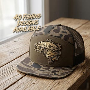 Op de afbeelding: Een camouflage truckerpet met een platte klep en een achterkant van gaas, voorzien van een centrale patch met een gedetailleerde illustratie van een baars en een berglandschap. De tekst 40 FISHING DESIGNS AVAILABLE staat linksboven.