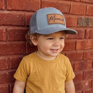 May include: A gray trucker hat featuring a rectangular tan leather patch on the front panel. The patch is embossed with the name Maverick in a black cursive script. The hat has a structured front and a mesh back with a curved brim.
