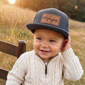 May include: A charcoal gray corduroy baseball cap featuring a rectangular tan leather patch on the front. The patch is embossed with the name Hayes in a black cursive script. The cap has a structured crown and a flat brim, providing a clean look.