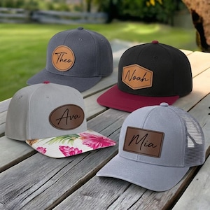 Peut inclure: Quatre casquettes de baseball avec des écussons en cuir personnalisés. Une casquette gris foncé avec un écusson rond portant le nom Theo, une noire à visière bordeaux avec un écusson hexagonal Noah, une gris clair à visière florale avec un écusson ovale Ava, et une casquette gris clair à filet avec un écusson rectangulaire Mia.