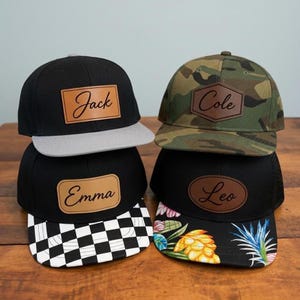 May include: Four baseball caps with leather-like patches featuring names. Top left: black cap with grey brim and rectangular patch reading Jack. Top right: camouflage cap with hexagonal patch reading Cole. Bottom left: black cap with chequered brim and rectangular patch reading Emma. Bottom right: black cap with floral brim and oval patch reading Leo.