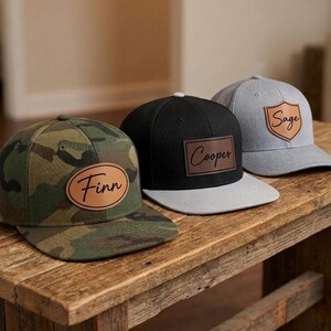 May include: Three baseball caps with leather patches. The first is camouflage with an oval patch reading Finn. The second is black with a rectangular patch reading Cooper. The third is light gray with a shield-shaped patch reading Sage.