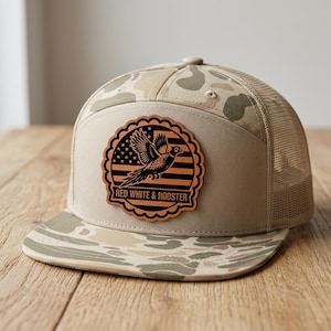 Op de afbeelding: Een beige honkbalpet met een camouflagepatroon op de klep en bovenpanelen, voorzien van een mesh achterkant. Een ronde leren patch op de voorkant toont het silhouet van een vliegende haan boven een gestreept vlagontwerp met de tekst RED WHITE & ROOSTER.