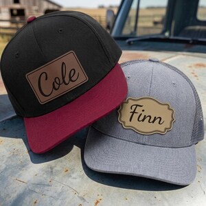 May include: A black baseball cap with a maroon brim and a rectangular brown leather patch engraved with the name Cole, alongside a grey mesh-back baseball cap featuring a tan scalloped leather patch engraved with the name Finn in black script.