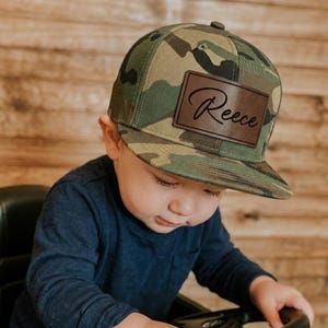 Pode incluir: Um boné de beisebol com estampa de camuflagem e um emblema retangular de couro marrom na parte frontal. O emblema tem o nome Reece gravado em letra cursiva preta. O boné possui uma copa estruturada e uma aba reta, mantendo um design clássico.