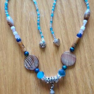 Op de afbeelding: Een handgemaakte ketting met blauwe, bruine en witte kralen, schelpvormige hangers en een kleine schelpenbedel. De ketting wordt gepresenteerd op een houten oppervlak.