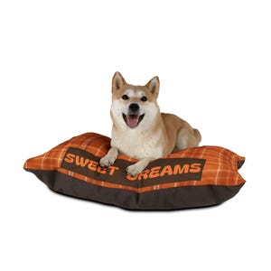 May include: A Shiba Inu dog rests on a rectangular dog bed. The bed has an orange plaid pattern with brown trim and the words "SWEET DREAMS" printed on it. The dog is light brown and white with its tongue out.