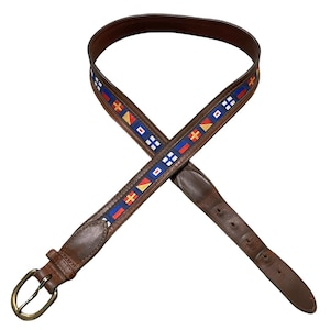 May include: A brown leather belt featuring a central ribbon strip decorated with a repeating pattern of colorful nautical signal flags in red, white, blue, and yellow. The belt includes a brass-toned metal buckle and multiple adjustment holes.