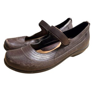 May include: A pair of brown leather Mary Jane style shoes. The shoes have a rounded toe, a strap across the top with a buckle closure, and a low heel. The leather has a worn appearance.