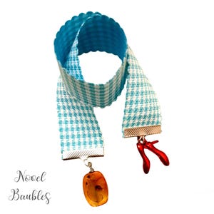 May include: A blue and white gingham bookmark with a scalloped edge. The bookmark has silver-toned metal ends, and is adorned with a red shoe charm and a pendant of amber-colored stone. The text "Novel Baubles" is in the lower left corner.